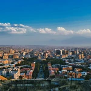 Veduta aerea di una città con un viale centrale, edifici e montagne sullo sfondo sotto un cielo blu e nuvoloso.