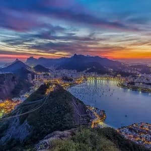 A panoramic view of an illuminated coastal city nestled between mountains and a bay under a colorful sunset sky.