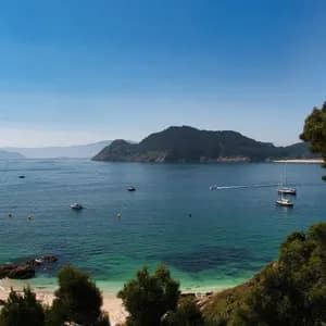 Una vista aérea de una bahía soleada con agua turquesa, una pequeña playa de arena y varios barcos navegando cerca de una isla boscosa.