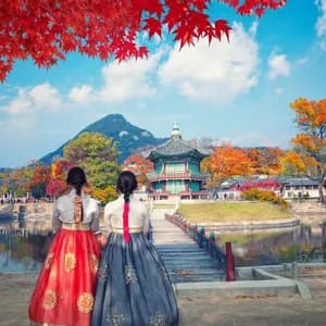 Due donne in abiti tradizionali hanbok guardano un lago verso una pagoda circondata da un vibrante fogliame autunnale.
