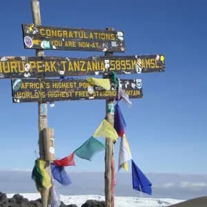 The wooden sign at Uhuru Peak, Tanzania, marking it as Africa's highest point, stands against a clear blue sky above the clouds.