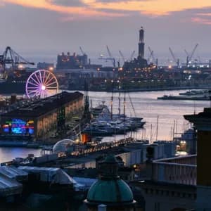 Una vista aerea di un porto al crepuscolo, che mostra una ruota panoramica illuminata, un faro, barche ormeggiate e gru industriali.