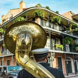 A musician from behind carries a large brass tuba on a street in front of a building with ornate, plant-filled balconies.