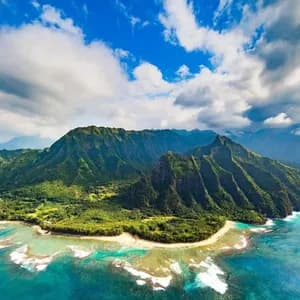 Veduta aerea di una costa montuosa e lussureggiante che incontra un oceano turchese, con spiagge sabbiose sotto un cielo azzurro nuvoloso.