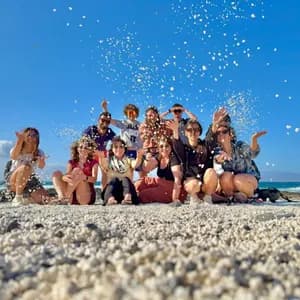 Un viaggio di gruppo WeRoad lancia sassolini bianchi in aria mentre posa per una foto su una spiaggia assolata.