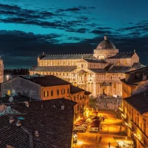 Veduta dall'alto di Torre Pendente, cattedrale e battistero illuminati al crepuscolo, sopra i tetti di una città storica.