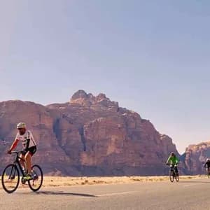 Un viaggio di gruppo WeRoad con persone in bicicletta su una strada asfaltata attraverso un paesaggio desertico con grandi formazioni rocciose sullo sfondo.