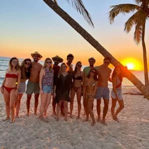Un grupo de WeRoad posa en una playa de arena con palmeras al atardecer sobre el océano.