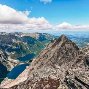 A panoramic view from a rocky mountain peak overlooking a vast mountain range with two blue alpine lakes in a valley below.