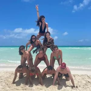 Un viaje en grupo de WeRoad forma una pirámide humana en una playa de arena frente a un mar turquesa bajo un cielo azul claro.