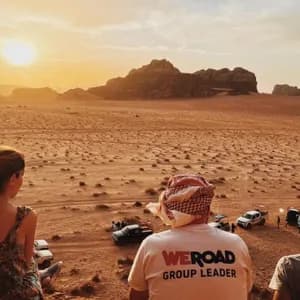 Un viaggio di gruppo WeRoad ammira il tramonto su un vasto paesaggio desertico con formazioni rocciose distanti e diverse jeep.