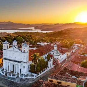 Eine Luftaufnahme einer weißen Kolonialkirche und einer Stadt mit roten Ziegeldächern, mit Blick auf einen See und Berge bei Sonnenuntergang.