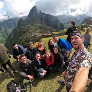 Un groupe WeRoad prend un selfie souriant sur un point de vue herbeux, surplombant des ruines antiques nichées dans des montagnes escarpées et verdoyantes.
