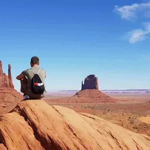 A person wearing a WeRoad backpack sits on a red rock, overlooking a desert valley with large buttes under a blue sky.