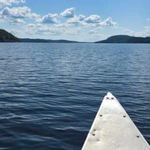 Eine Ich-Perspektive von einem Kanu auf einem großen See, mit bewaldeten Hügeln in der Ferne unter blauem Himmel mit weißen Wolken.