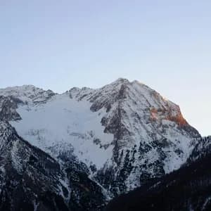 A snow-covered mountain peak is illuminated by the orange light of sunrise under a clear sky, with dark forested slopes below.