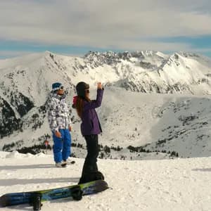 Dos personas con equipo de nieve están de pie en la cima de una montaña nevada, mientras una de ellas fotografía la inmensa cordillera. Una tabla de snowboard descansa en primer plano.