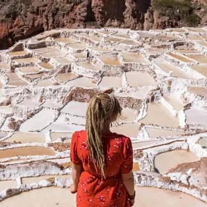 A woman with blonde dreadlocks in a red top, seen from behind, looking out over terraced salt evaporation ponds.