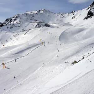 Una vista ampia di una pista da sci affollata con molte persone che sciano, circondata da montagne innevate e impianti di risalita.