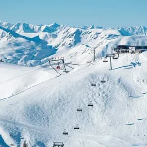A chairlift ascends a snow-covered mountain slope toward a summit station, with a panoramic view of a snowy mountain range.