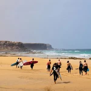 Un groupe WeRoad en combinaisons de surf, planches à la main, marche sur une plage de sable vers l'océan, avec des falaises au loin.