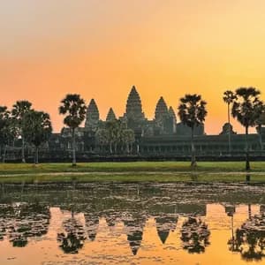 An ancient temple with tall spires and palm trees reflected in a calm body of water against an orange sky at sunrise.