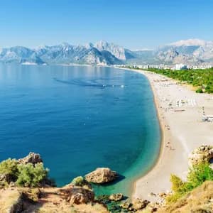 A panoramic view of a long sandy beach along a calm blue sea, with a coastal city and mountains in the background.