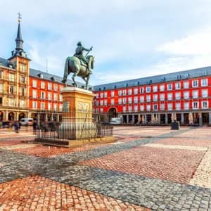 A bronze equestrian statue stands in the center of a large cobblestone plaza, surrounded by historic red buildings as people walk by.