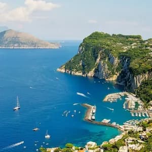 Ein erhöhter Blick auf den Yachthafen einer Küstenstadt, gefüllt mit Booten, eingebettet neben einer grünen, felsigen Klippe unter blauem Himmel.