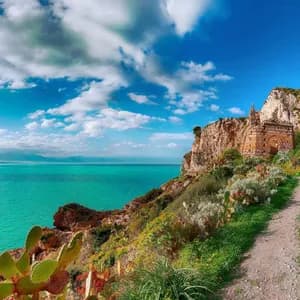 Un chemin de terre mène à un monument en pierre sur une falaise verdoyante surplombant une mer turquoise sous un ciel nuageux.