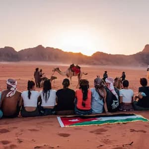 Un groupe WeRoad assis sur une dune de sable rouge, avec un drapeau jordanien, admirant le coucher de soleil sur le désert, avec des montagnes et des chameaux en arrière-plan.