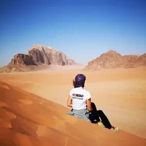 Un capogruppo WeRoad visto di spalle, seduto su una vasta duna di sabbia arancione e che guarda montagne rocciose lontane sotto un cielo azzurro chiaro.