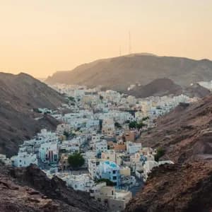 A town of white buildings nestled in a valley surrounded by rocky mountains at sunset.