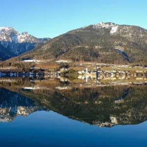 Un borgo lacustre e montagne innevate che si riflettono perfettamente nelle acque calme sotto un cielo azzurro.