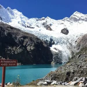 Un cartello di legno per Arhuaycocha accanto a un lago glaciale turchese ai piedi di imponenti montagne innevate e un ghiacciaio.
