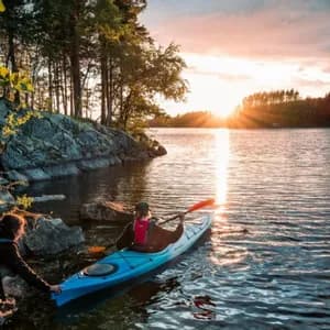 Eine Person an einem felsigen Ufer hilft einer anderen Person in einem blauen Kajak, bei Sonnenuntergang in einen See zu starten.