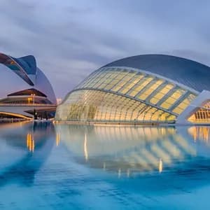 Dos edificios modernos e iluminados con arquitectura curva reflejándose en un gran y tranquilo espejo de agua al anochecer.