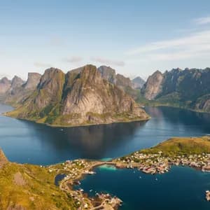 Una vista aerea di un villaggio costiero ai piedi di grandi montagne rocciose circondato da un fiordo blu profondo.
