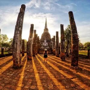 Una persona si erge tra antichi pilastri di fronte a una statua di Buddha e a uno stupa, con lunghe ombre proiettate dal sole al tramonto.
