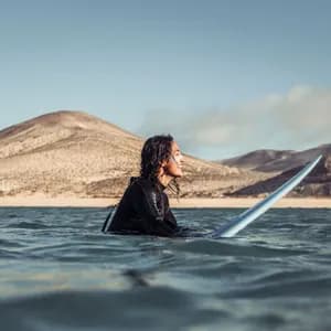 Una persona con i capelli bagnati in una muta nera sorride mentre è seduta su una tavola da surf nell'oceano, con colline sabbiose sullo sfondo.