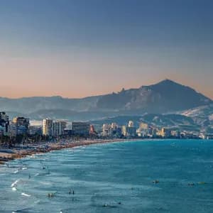 Un paesaggio urbano su una costa con una spiaggia affollata dove le persone nuotano nell'oceano, con grandi montagne visibili in lontananza.