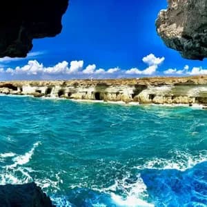 Vue de l'intérieur d'une grotte marine donnant sur un littoral rocheux avec d'autres grottes marines, un océan turquoise et un ciel bleu nuageux.