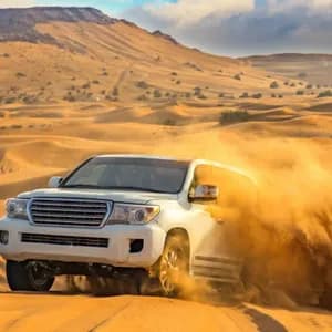 Un SUV bianco solleva una nuvola di sabbia mentre attraversa un vasto deserto con grandi dune sullo sfondo.
