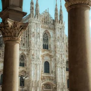 The ornate white facade of a Gothic cathedral with numerous spires, framed between two large, carved stone columns.