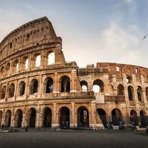 El antiguo anfiteatro Coliseo en Roma, visto desde una plaza de adoquines vacía al amanecer bajo un cielo parcialmente nublado.