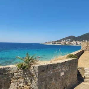 Une vue depuis un mur de pierre surplombant une ville côtière sur une baie aux eaux turquoise et aux collines verdoyantes sous un ciel clair.