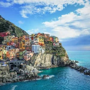 A village of colorful houses built on a rocky cliffside, overlooking a calm blue sea under a partly cloudy sky.