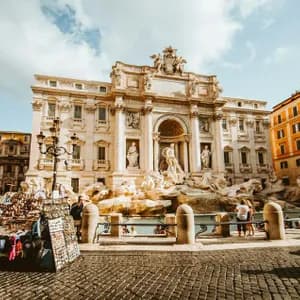 An ornate white marble fountain in a city square on a sunny day, with a souvenir stall on the cobblestone foreground.