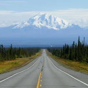 A straight paved road stretches through a forest of evergreens towards a large, snow-capped mountain range in the distance.
