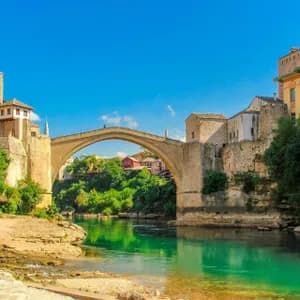 Un ponte ad arco in pietra attraversa un fiume turchese, fiancheggiato da edifici storici e una vegetazione lussureggiante, sullo sfondo di un cielo azzurro luminoso.